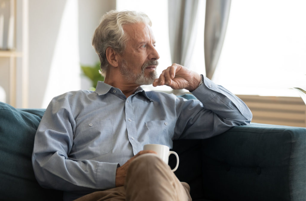 A senior man holding a cup of tea sitting on a couch and looking out the window with a serious expression.