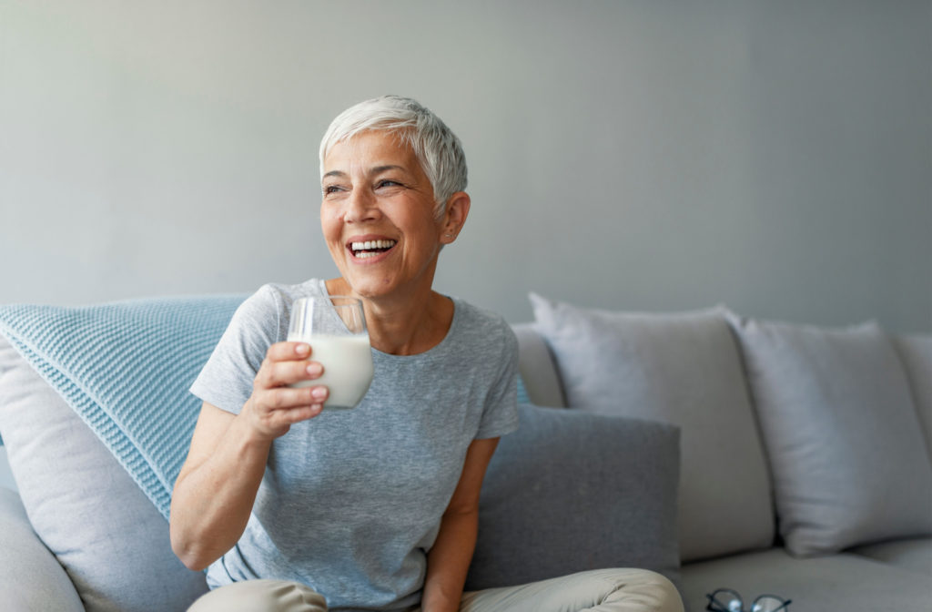 A an older woman smiling and sitting on a couch while holding a glass of milk on her right hand.