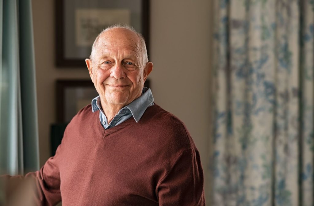 A smiling senior man looking out the window in his own home.