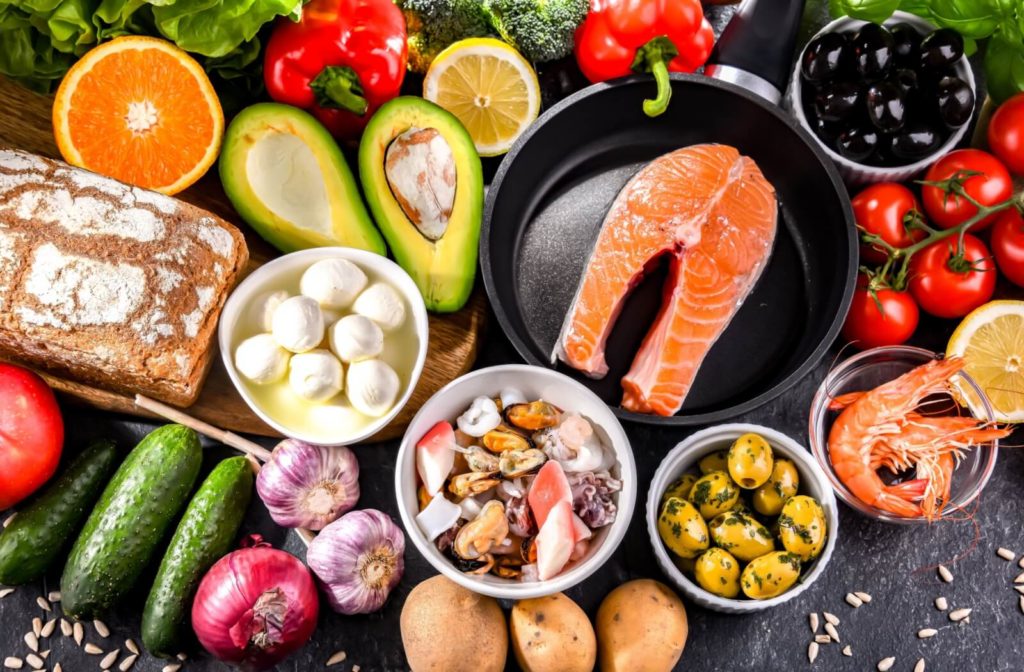 A variety of fruits, vegetables, nuts, meats, and bread used in the Mediterranean diet arranged on a black table.