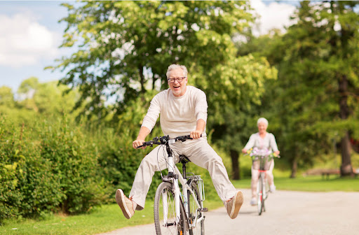 A senior man on a bike holds his feet off the pedals out to the side while smiling. a senior woman bikes in the background behind him.