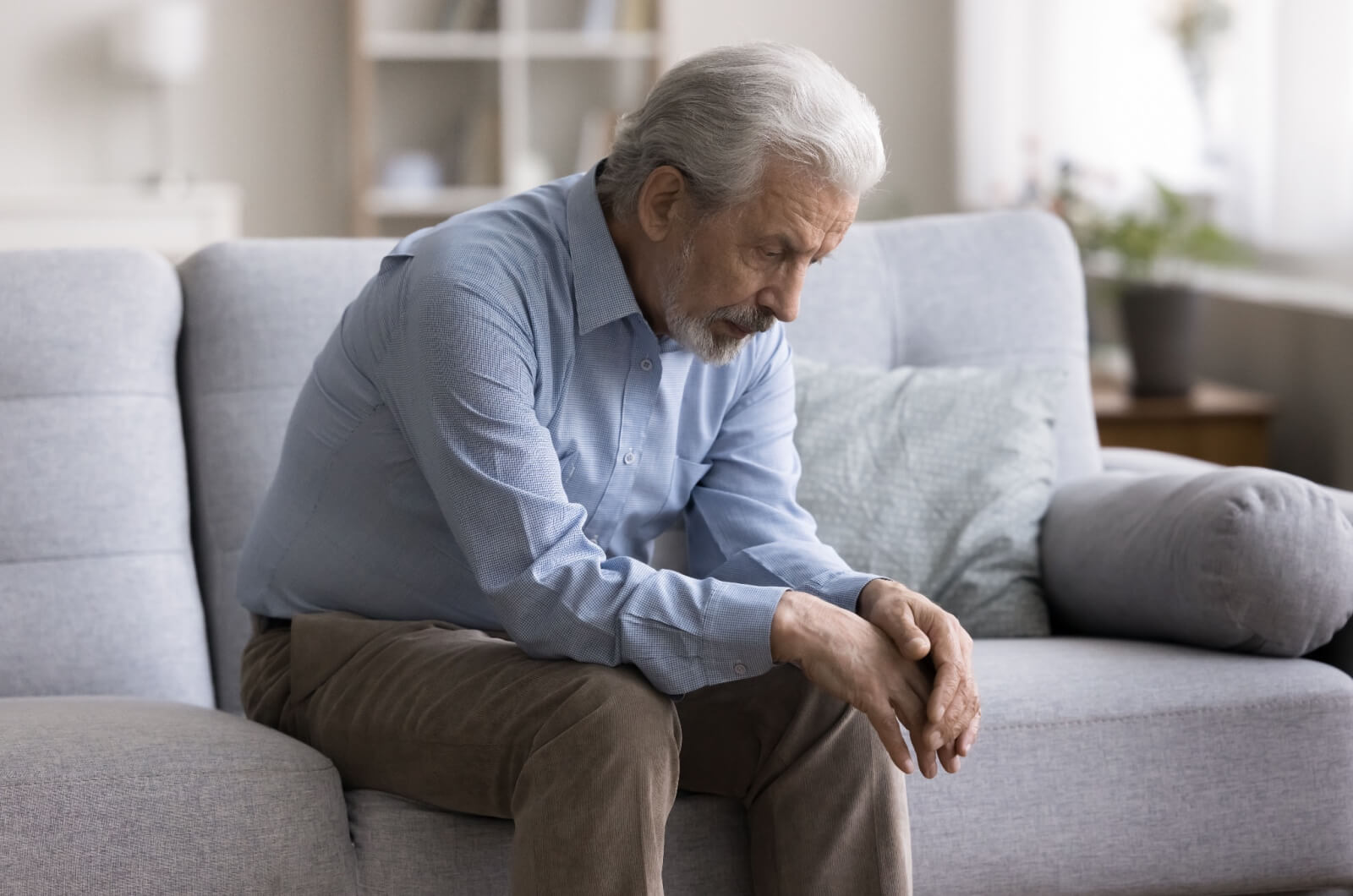 A senior with early signs of dementia sitting on a couch with head bowed and elbows on his knees.