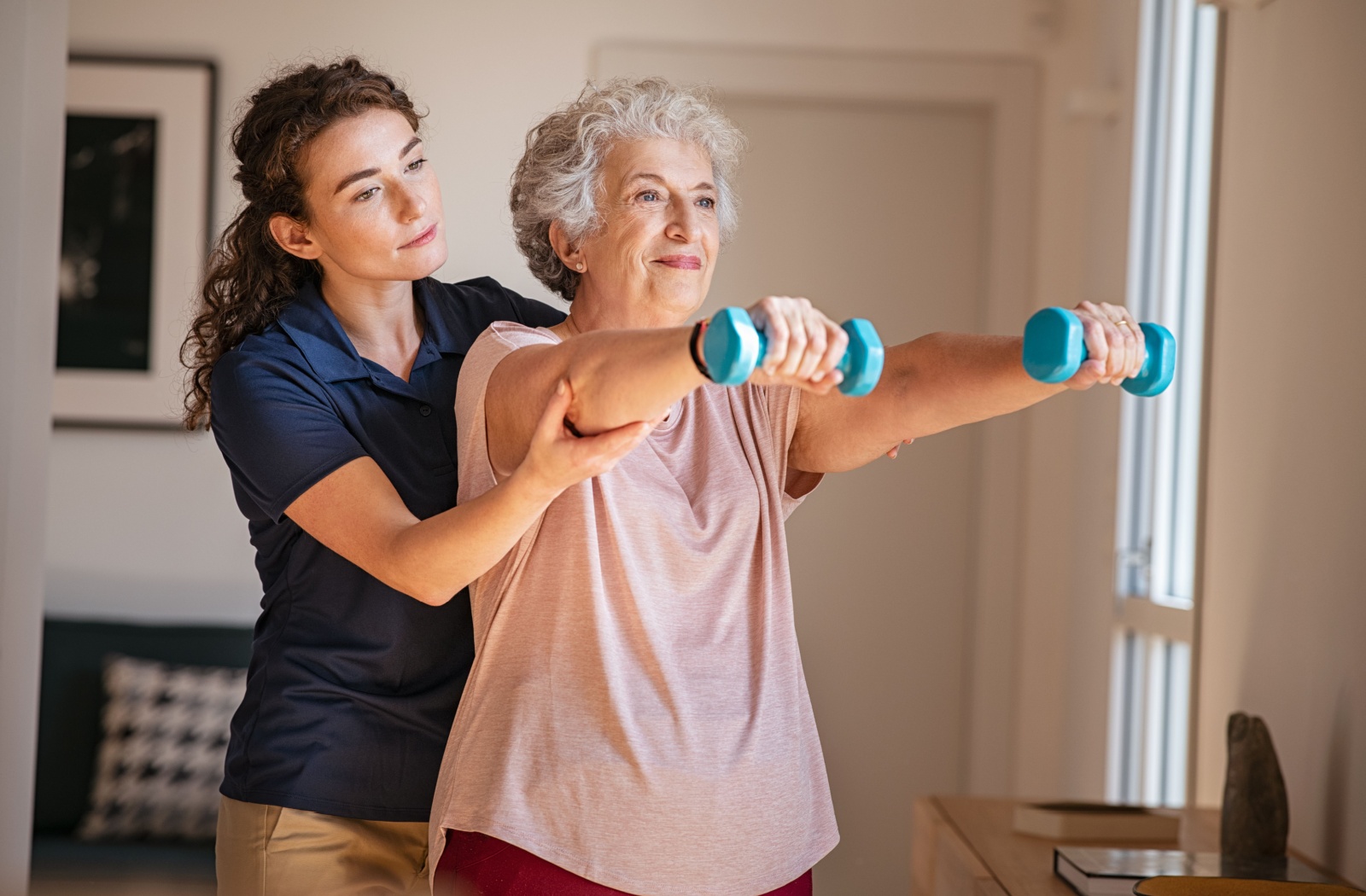 A caregiver in assisted living helping an older adult lift small weights during a physical therapy exercise.