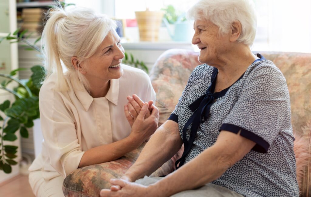 An older adult sits on a couch and chats with their caregiver who is visiting them during a stay in a respite care community.