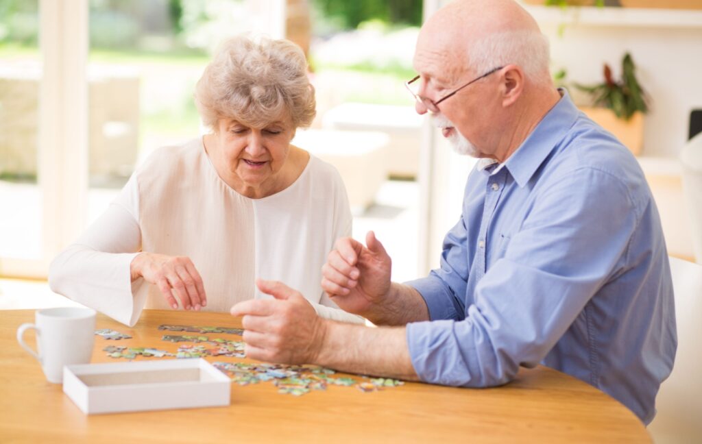 A senior couple sits at their kitchen table with a jigsaw puzzle in front of them. They chat while they fit puzzle pieces together.