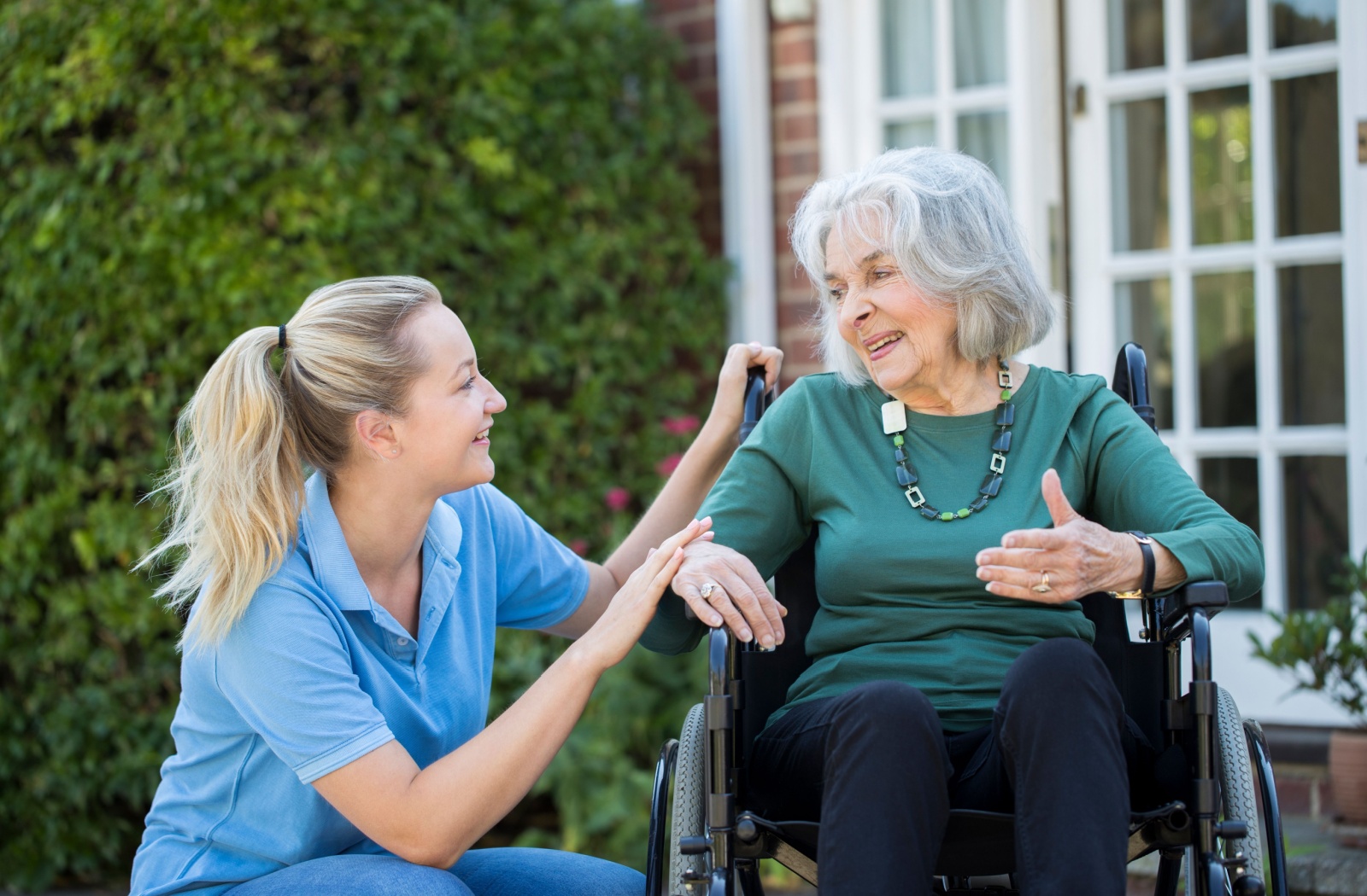 A friendly caregiver kneels down to talk to a dementia patient in a wheelchair at eye level to help them feel heard.