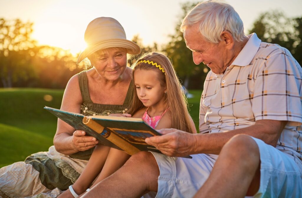 A senior couple sit outdoors with their grandchild, sharing memories in a photo album while the sun sets.
