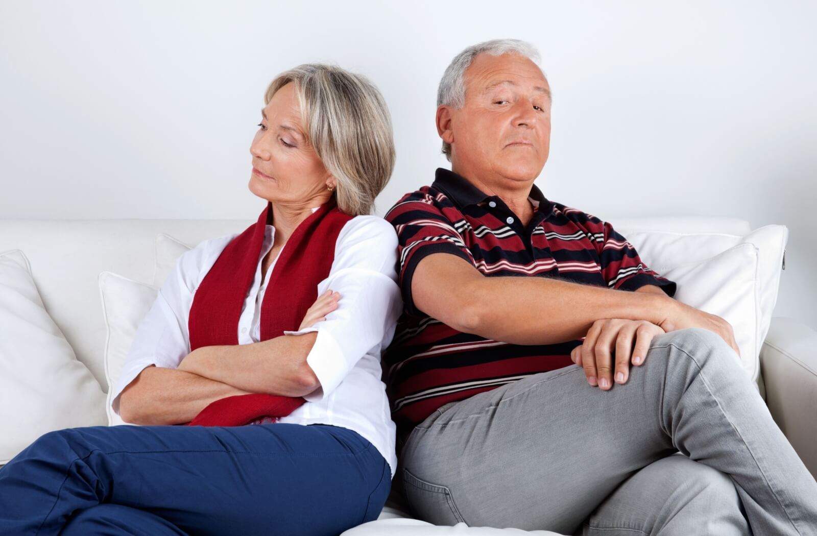 A senior couple sit back to back on a white sofa following an argument about their differing care needs
