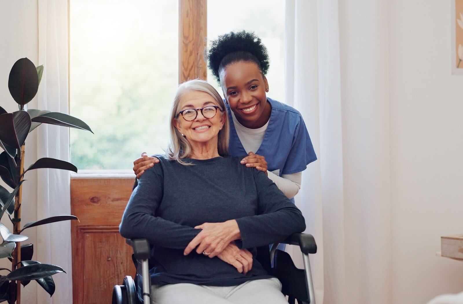 A nurse smiles behind an older adult, gently hugging them, showcasing a close relationship and level of trust