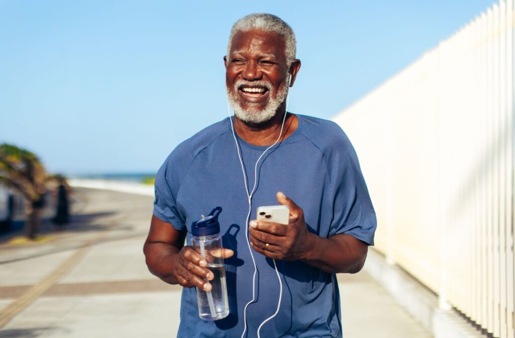 A happy senior goes for a morning run, holding a water bottle and smartphone, while wearing earphones and listening to music.