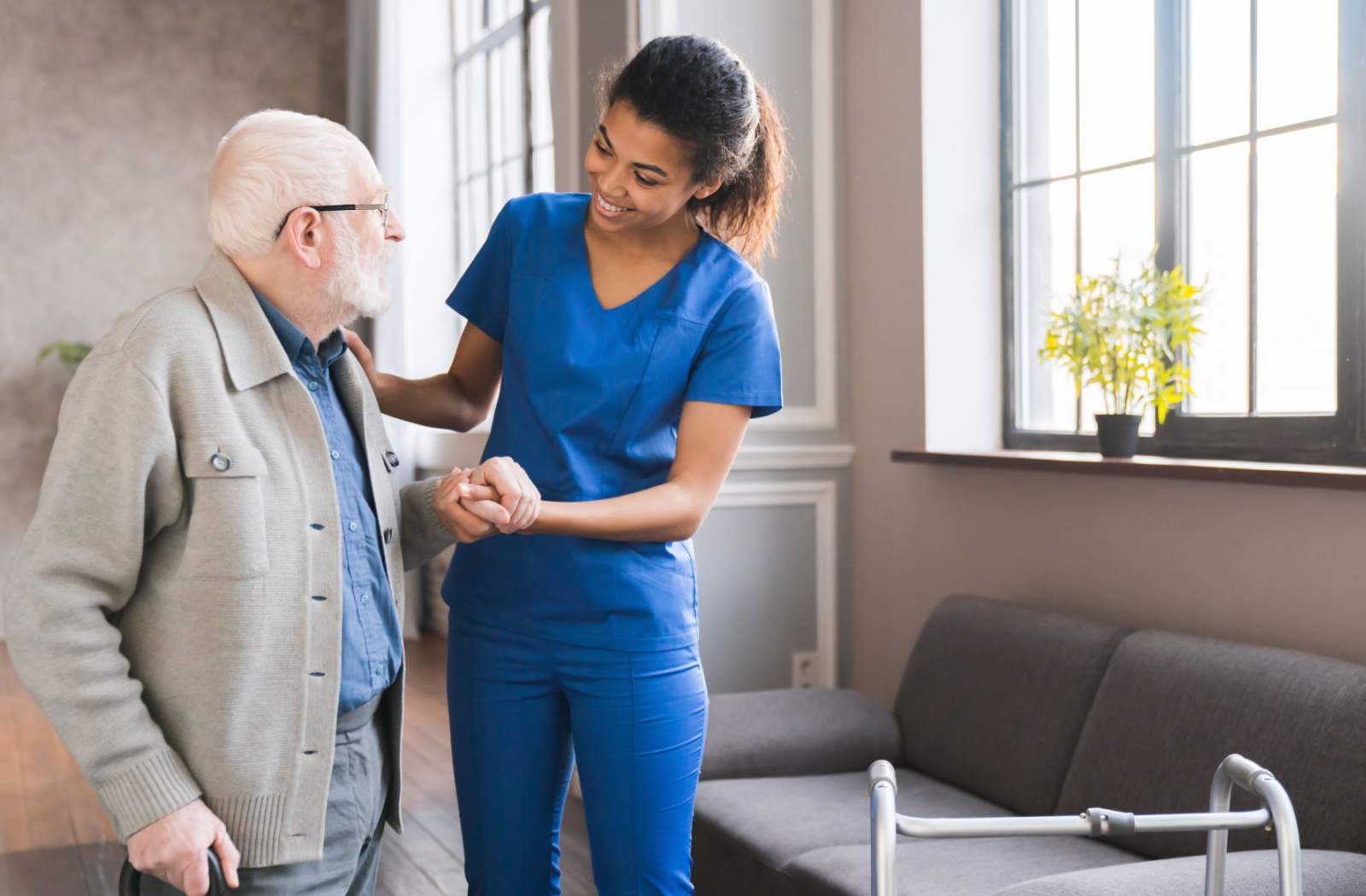 A smiling caregiver offers support for a senior walking with a cane in a bright respite care setting.