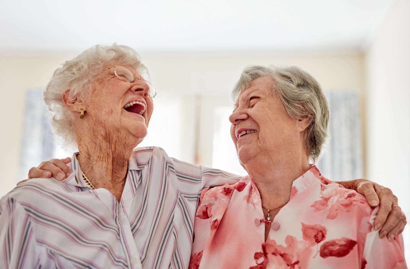 Two senior women laugh as they hold each other, enjoying each other's company.