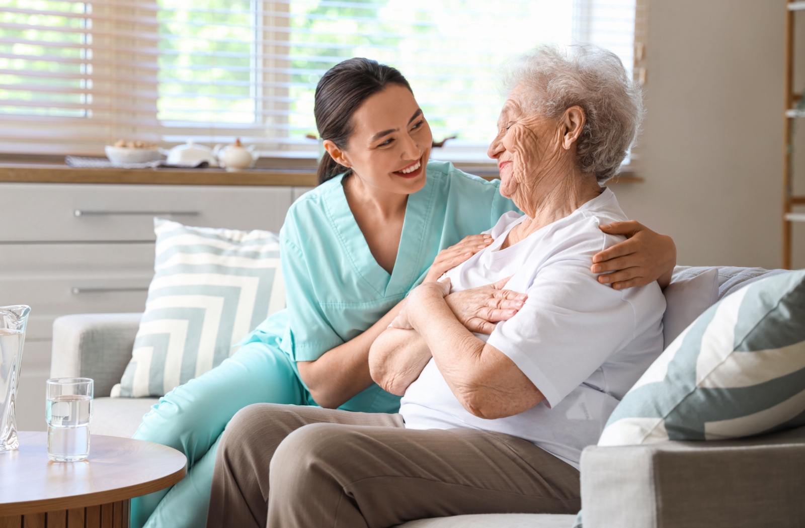 A smiling caregiver and senior sit next to each other on a sofa in a bright room in respite care.